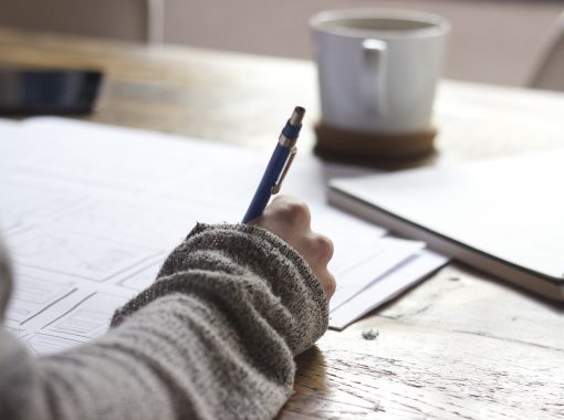 Picture of a woman writing on paper with a coffee cup in the background. Image used for blog about how to write a press release summary.