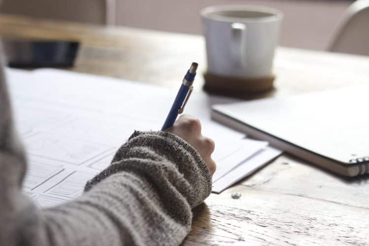 Picture of a woman writing on paper with a coffee cup in the background. Image used for blog about how to write a press release summary.
