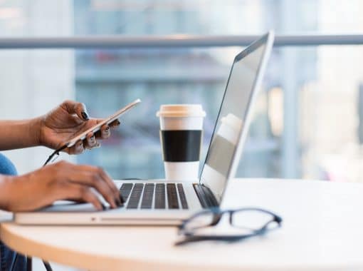 Picture of woman typing on computer, looking at phone, with coffee cup near by. Picture being used for blog, "Are Press Releases Driving Business Growth for CEOs?"