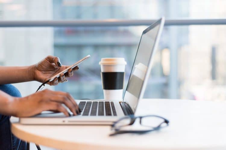 Picture of woman typing on computer, looking at phone, with coffee cup near by. Picture being used for blog, "Are Press Releases Driving Business Growth for CEOs?"