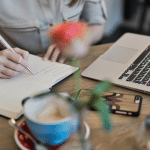 Image of a lady writing in a notebook on a table with a phone, blue coffee cup, and Apple laptop. Image is being used for a Newswire blog about sponsored content.