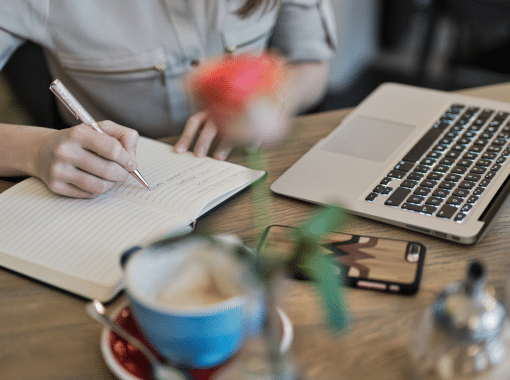 Image of a lady writing in a notebook on a table with a phone, blue coffee cup, and Apple laptop. Image is being used for a Newswire blog about sponsored content.