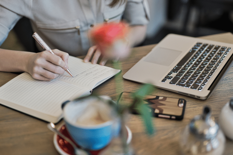 Image of a lady writing in a notebook on a table with a phone, blue coffee cup, and Apple laptop. Image is being used for a Newswire blog about sponsored content.