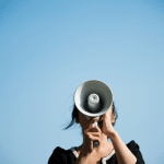 Image of a lady speaking into a megaphone with a blue sky background. Picture is being used for a blog post about press release campaigns.