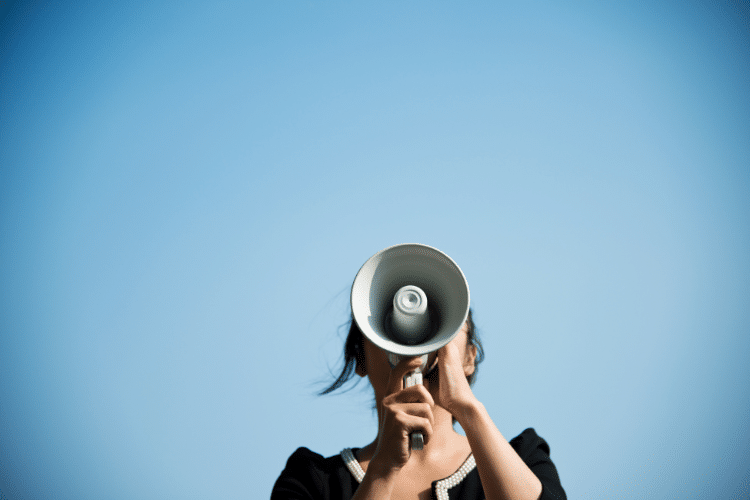 Image of a lady speaking into a megaphone with a blue sky background. Picture is being used for a blog post about press release campaigns.
