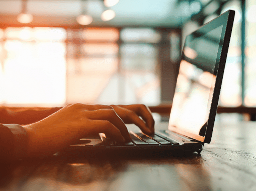 Picture of a person typing on a laptop. Computer is in the forefront, light in the background. Picture is being used for a blog post about how much does it cost to distribute a press release?