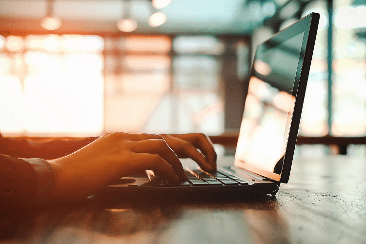 Picture of a person typing on a laptop. Computer is in the forefront, light in the background. Picture is being used for a blog post about how much does it cost to distribute a press release?