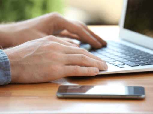 Image of a man's hands on the keyboard of a laptop. A smartphone is next to him. Image is being used for a Newswire blog title, Where do Press Releases Get Distributed?