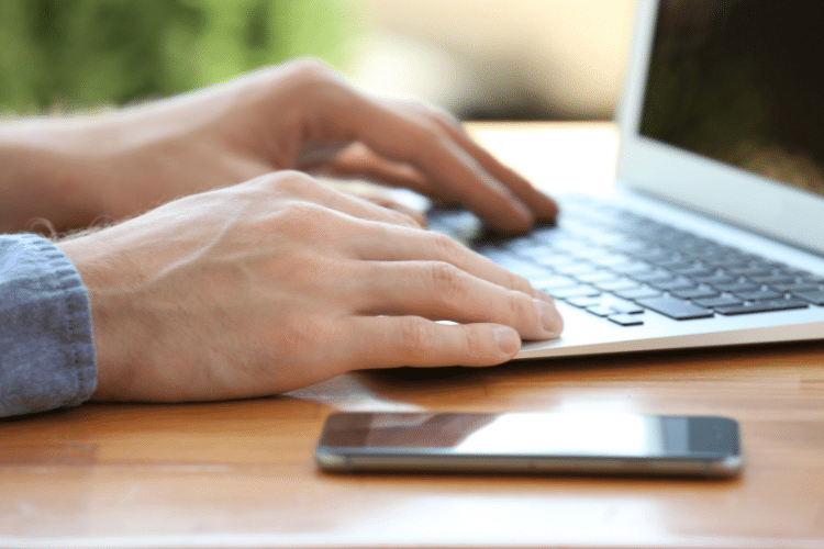 Image of a man's hands on the keyboard of a laptop. A smartphone is next to him. Image is being used for a Newswire blog title, Where do Press Releases Get Distributed?