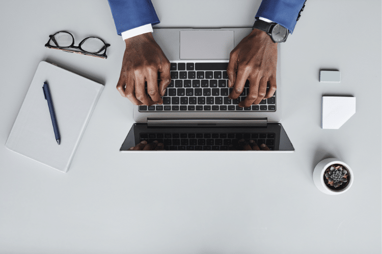 Flat lay image of an African American man in a blue suit typing on a Macbook. Image is being used for a blog about press release distribution outsourcing.