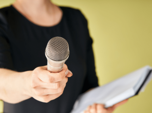 Picture of a lady in a black shirt holding a microphone out and a notebook on a yellow backdrop. This image is being used for a blog about preparing for a media interview.