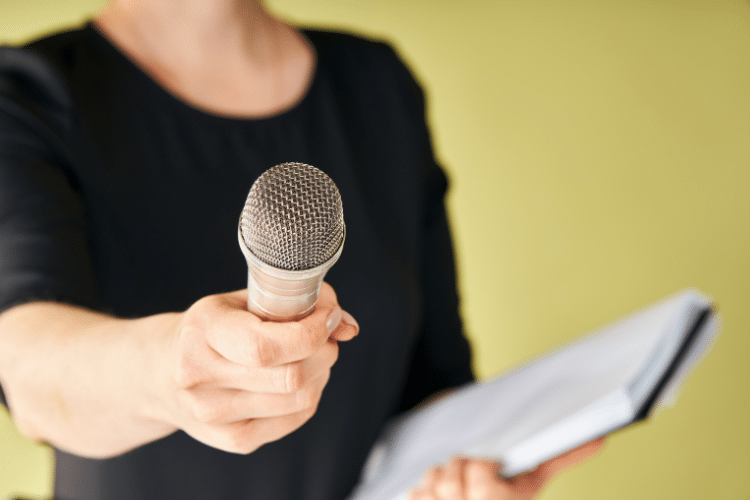 Picture of a lady in a black shirt holding a microphone out and a notebook on a yellow backdrop. This image is being used for a blog about preparing for a media interview.