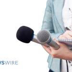 Newswire-branded image of a lady in a light blue blazer with a white button down, writing in a notebook and holding two microphones.