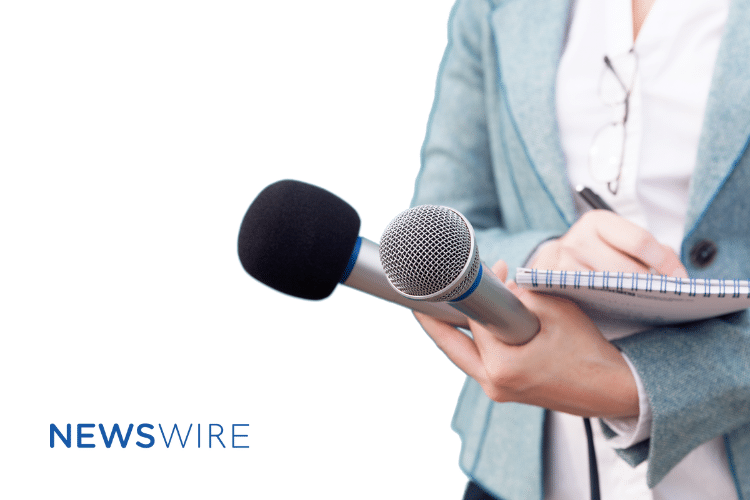 Newswire-branded image of a lady in a light blue blazer with a white button down, writing in a notebook and holding two microphones.