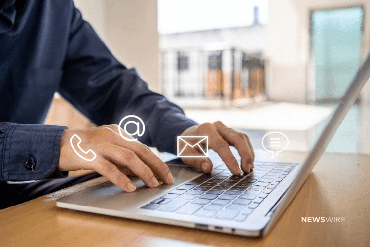 Picture of a man in a blue button down typing on a computer. There are four symbols near his hands, telephone symbol, ampersand, an envelope, and a speech bubble. Image is being used for a Newswire blog about a Media Database.