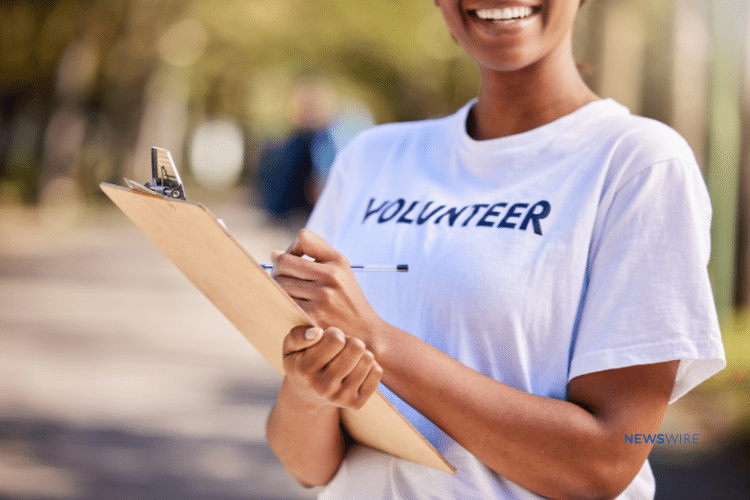 Picture of an African American woman holding a clipboard and smiling at a charity event. Her shirt says, "volunteer." This image is being used for a Newswire blog about How to Write a Nonprofit Press Release